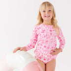 Child wearing a pink and white patterned long sleeve two-piece swimsuit on a white background holding a pink floatie
