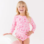 Child wearing a pink and white patterned long sleeve two-piece swimsuit on a white background, holding a pink floatie