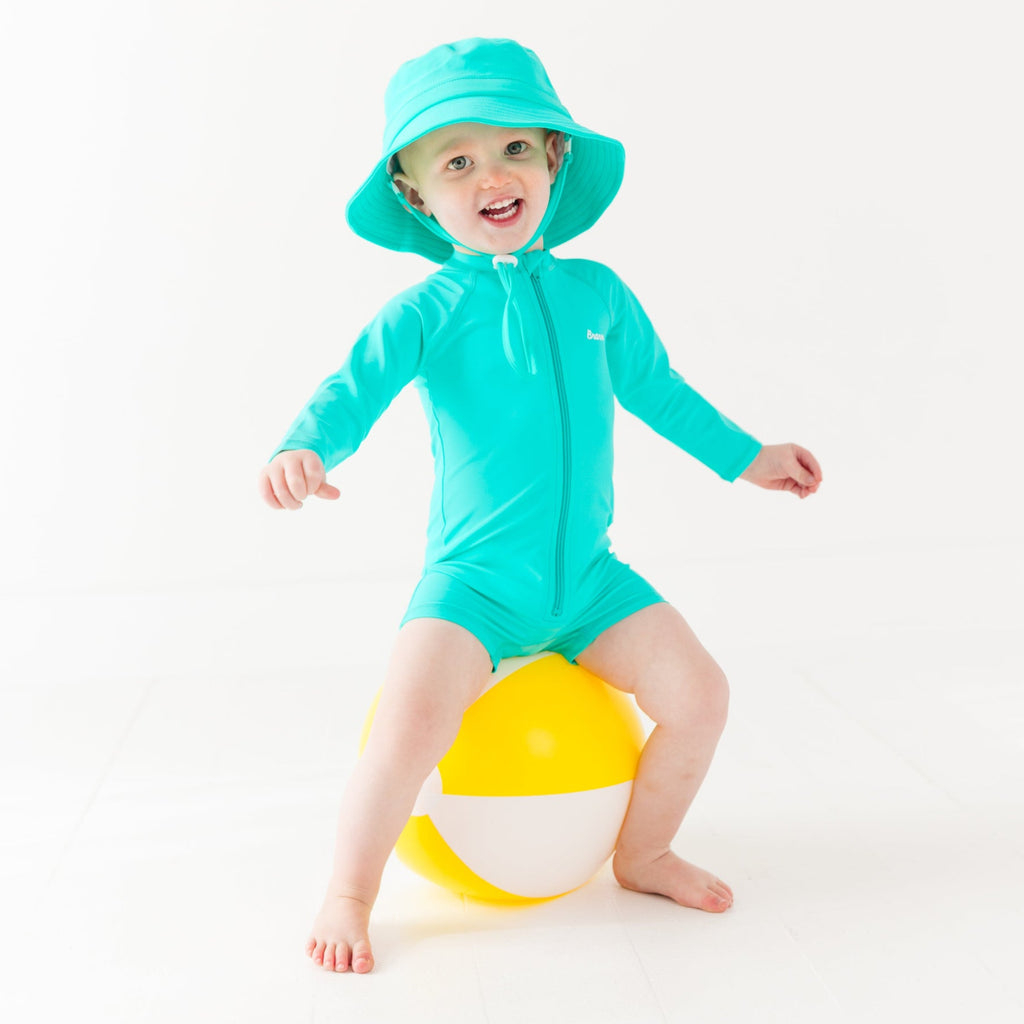 Child wearing a turquoise boy swimsuit and matching bucket hat with a yellow beach ball on a white background