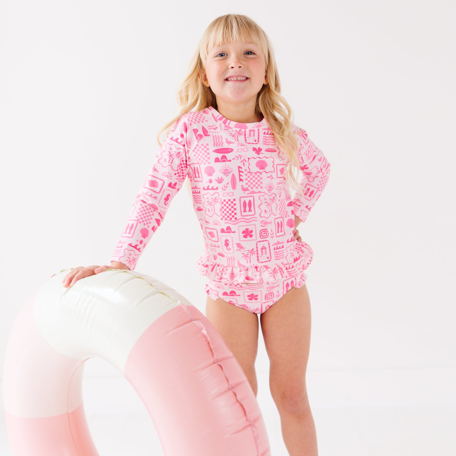 Child wearing a pink and white patterned long sleeve two-piece swimsuit on a white background holding a pink floatie