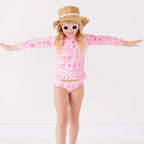 Child wearing a pink and white patterned long sleeve two-piece swimsuit, flower sunglasses and brown hat on a white background