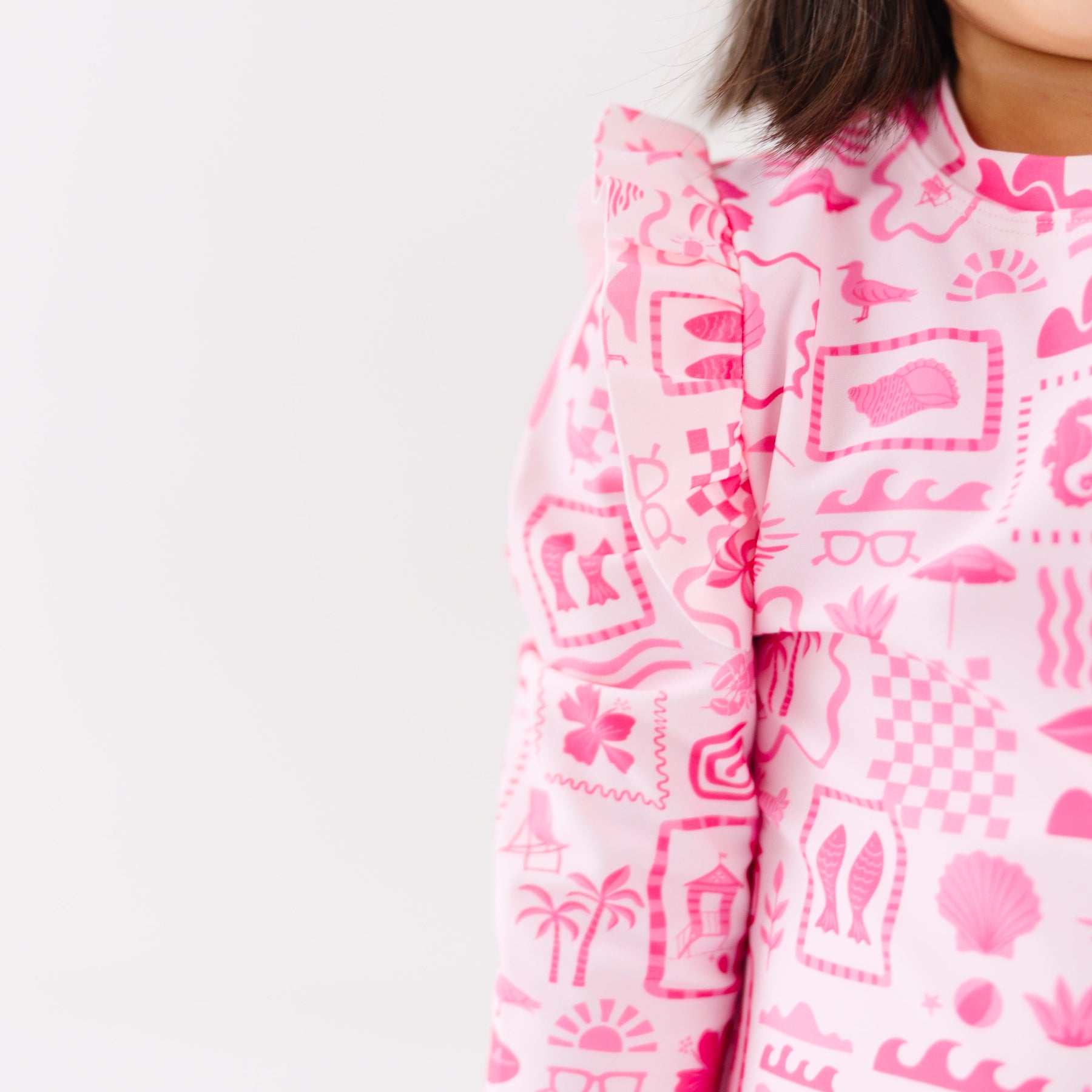 Close up shot of a child wearing a pink and white patterned long sleeve one-piece swimsuit and a matching light pink hat on a white background