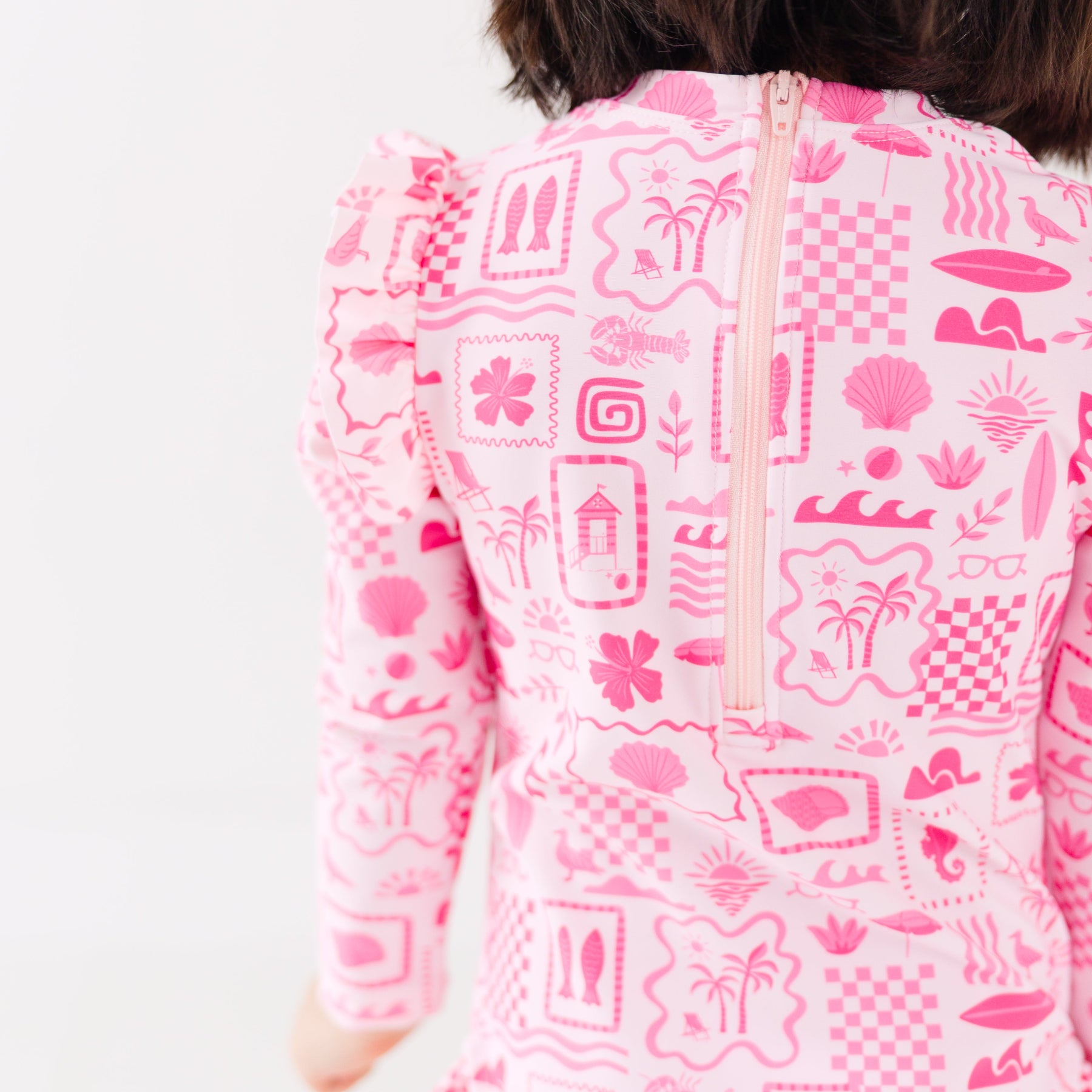 Back view of a child wearing a pink and white patterned long sleeve one-piece swimsuit on a white background