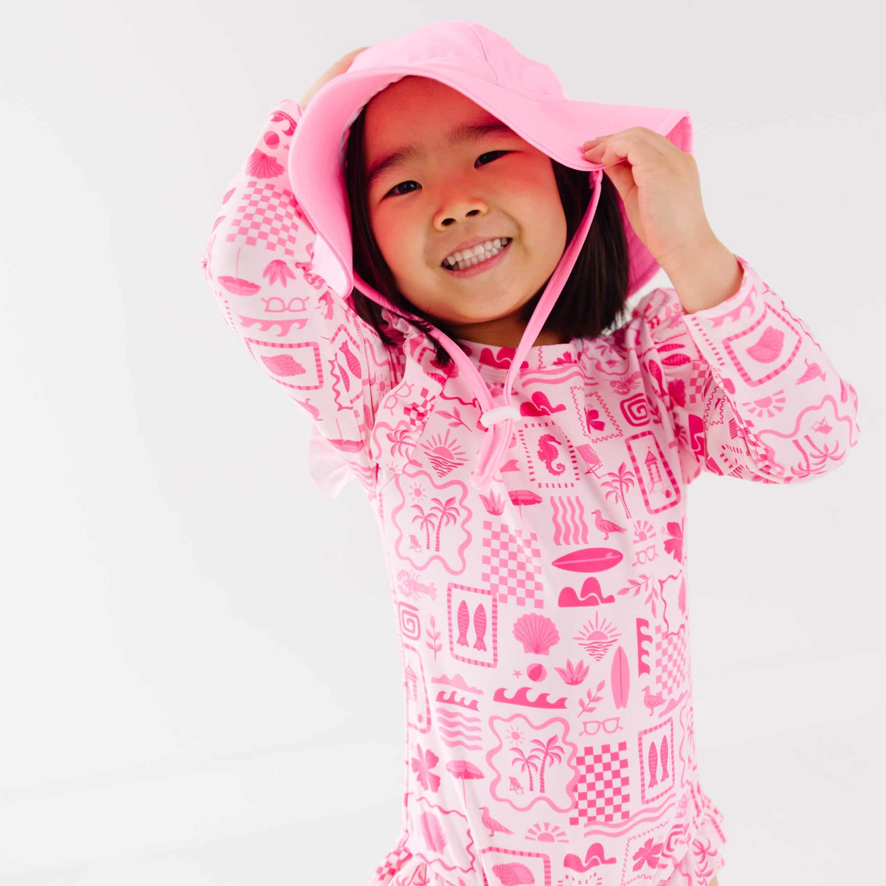 Child wearing a pink and white patterned long sleeve one-piece swimsuit and a matching light pink hat on a white background