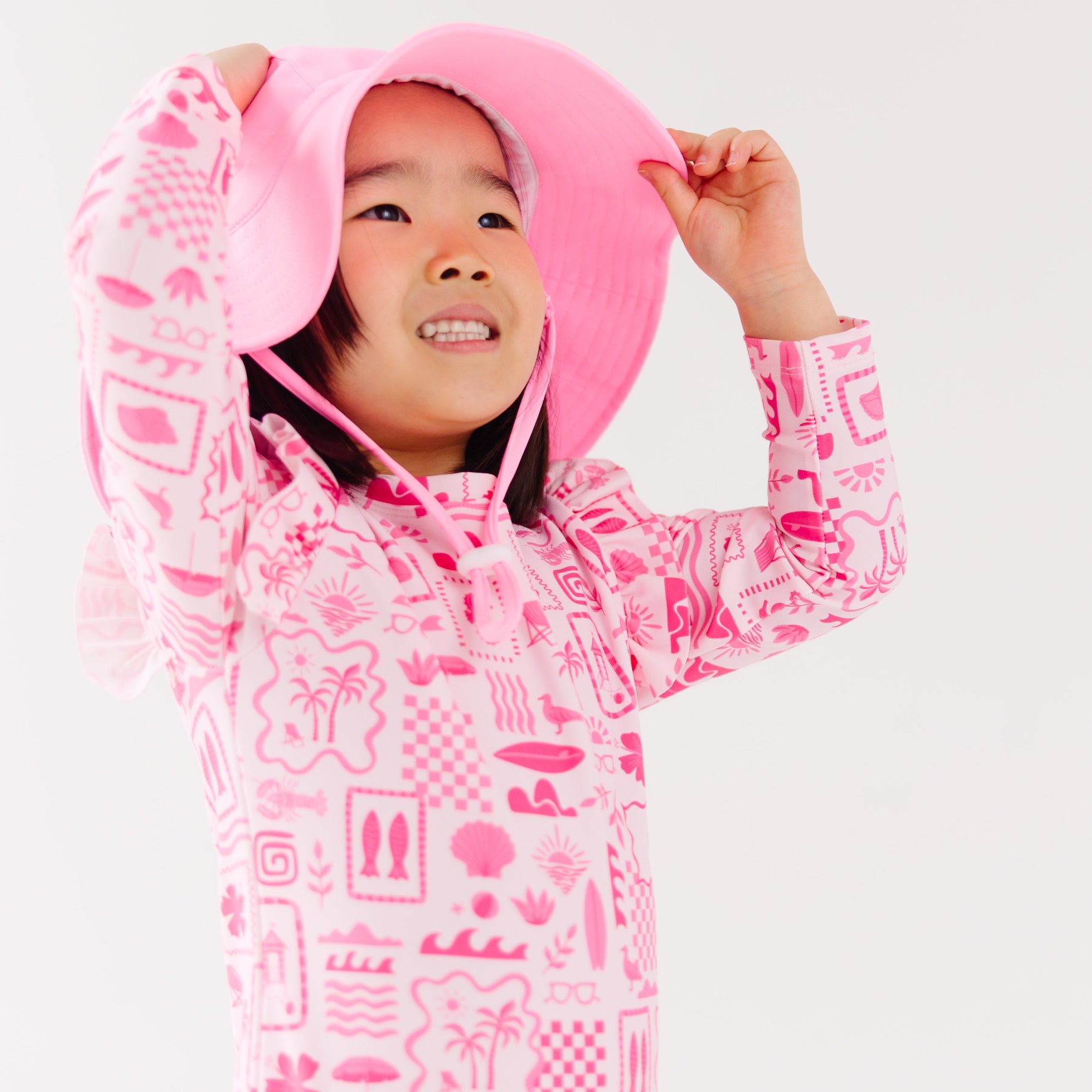 Child wearing a pink and white patterned long sleeve one-piece swimsuit and a matching light pink hat on a white background
