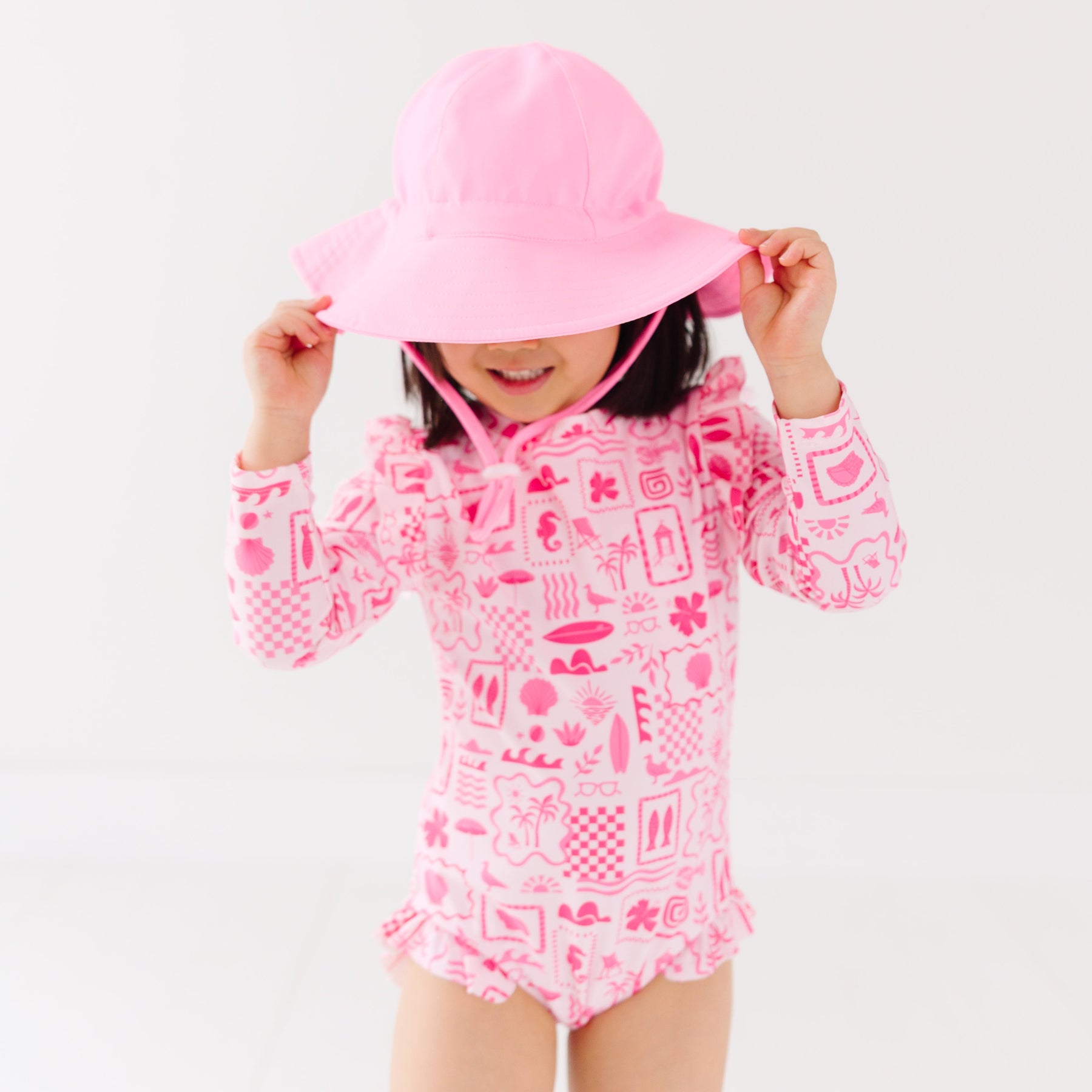 Child wearing a pink and white patterned long sleeve one-piece swimsuit and a matching light pink hat on a white background