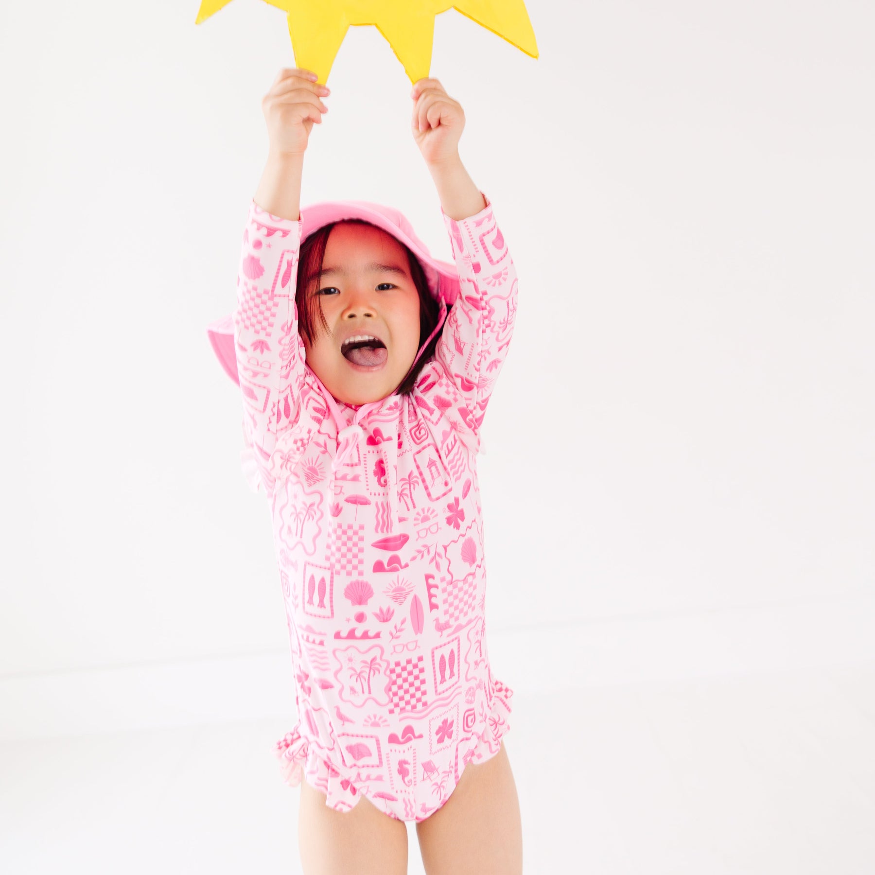 Child wearing a pink and white patterned long sleeve one-piece swimsuit and a matching light pink hat with a yellow star-shaped object above their head on a white background