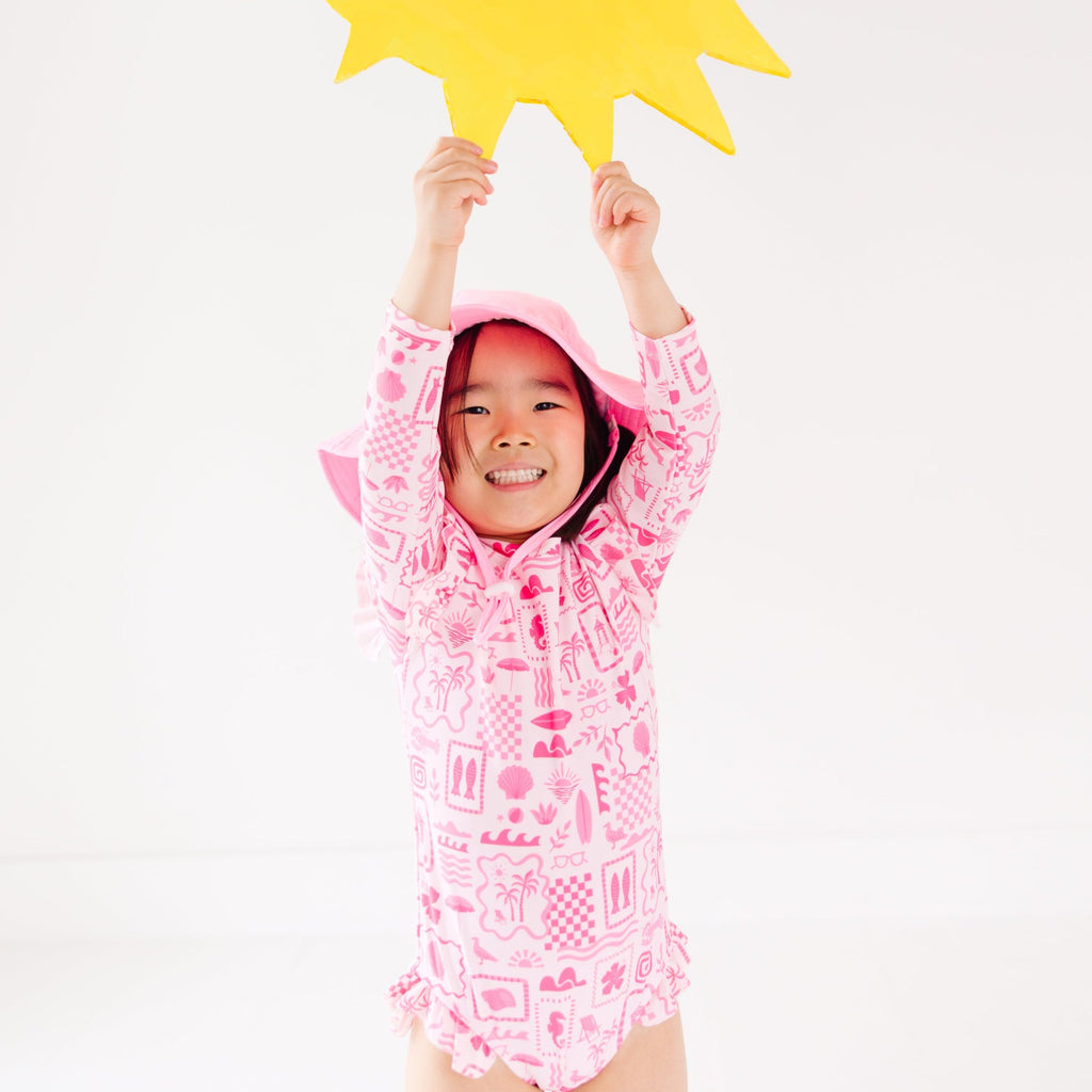 Child wearing a pink and white patterned long sleeve one-piece swimsuit and a matching light pink hat with a yellow star-shaped object above their head on a white background