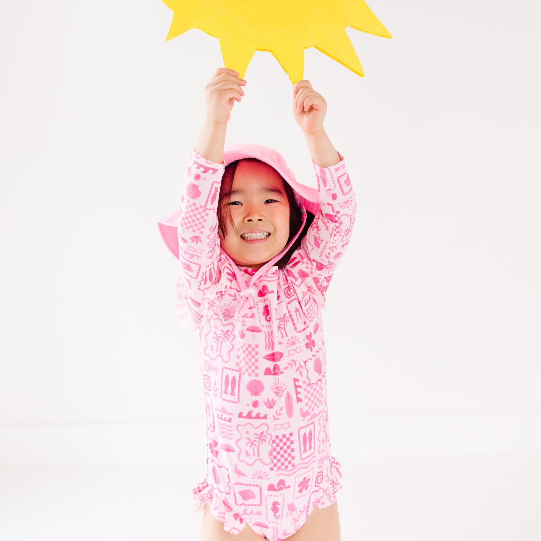 Child wearing a pink and white patterned long sleeve one-piece swimsuit and a matching light pink hat with a yellow star-shaped object above their head on a white background