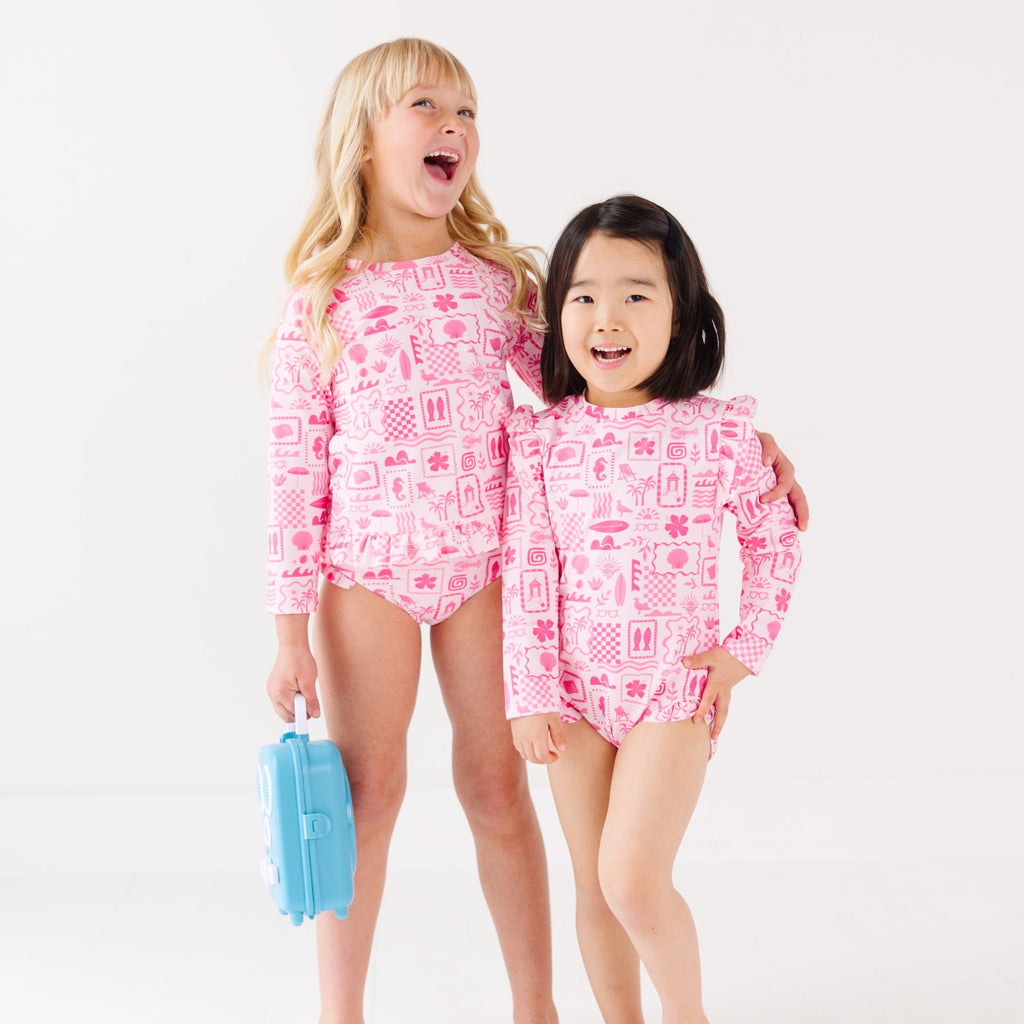Two children wearing a pink and white patterned long sleeve one-piece and two-piece swimsuit holding hands with a blue radio toy.