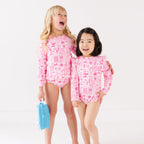 Two children wearing a pink and white patterned long sleeve one-piece and two-piece swimsuit holding hands with a blue radio toy.