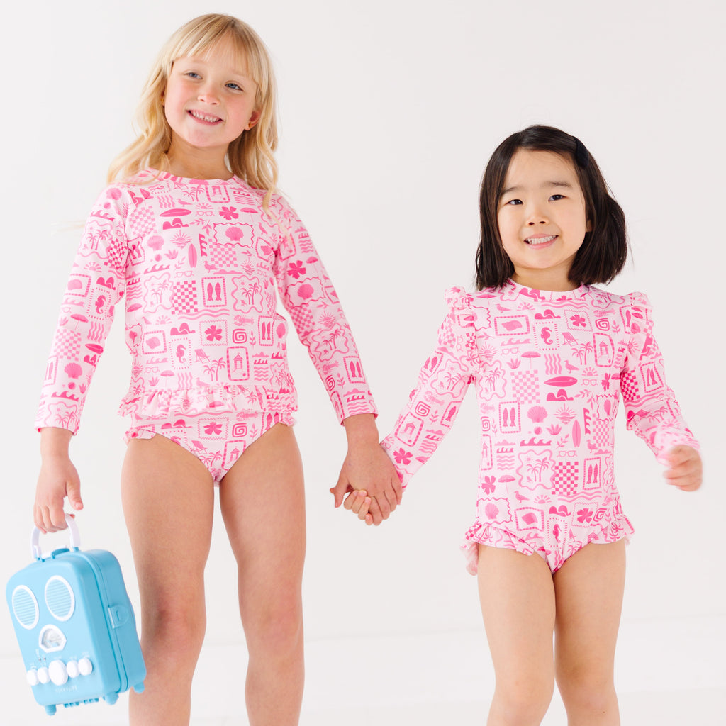 Two children wearing a pink and white patterned long sleeve one-piece and two-piece swimsuit holding hands with a blue radio toy.