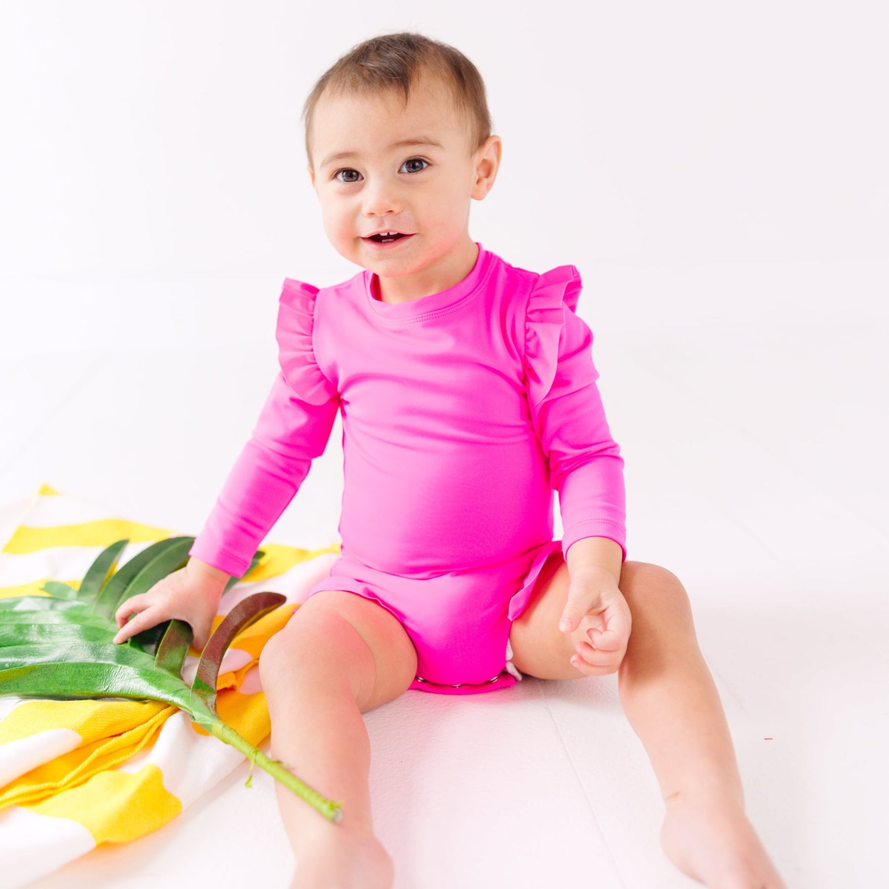 Baby wearing a bright pink one-piece swimsuit sitting on a white background with a yellow towel and green leaves.