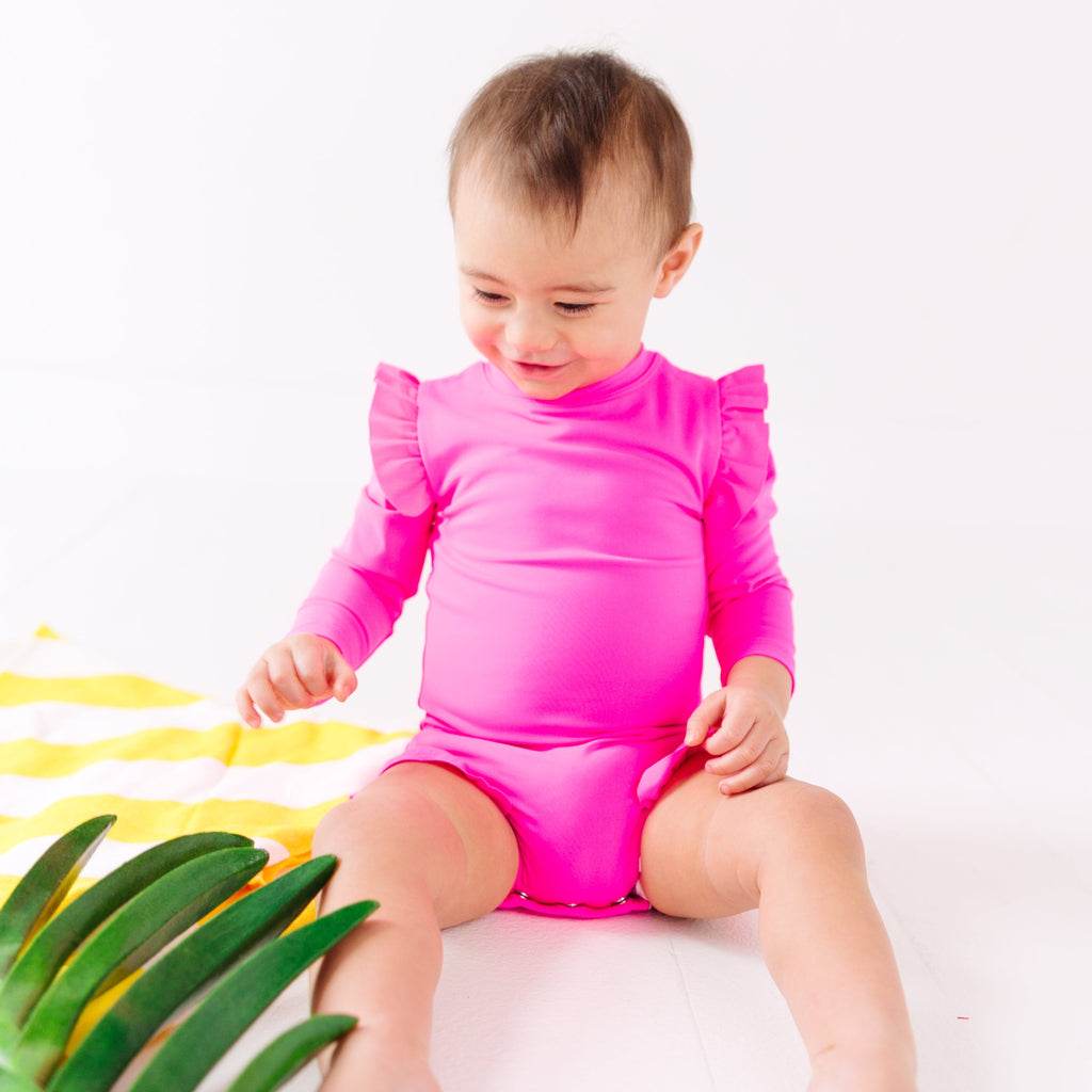 Baby wearing a bright pink one-piece swimsuit sitting on a white background with a yellow towel and green leaves.