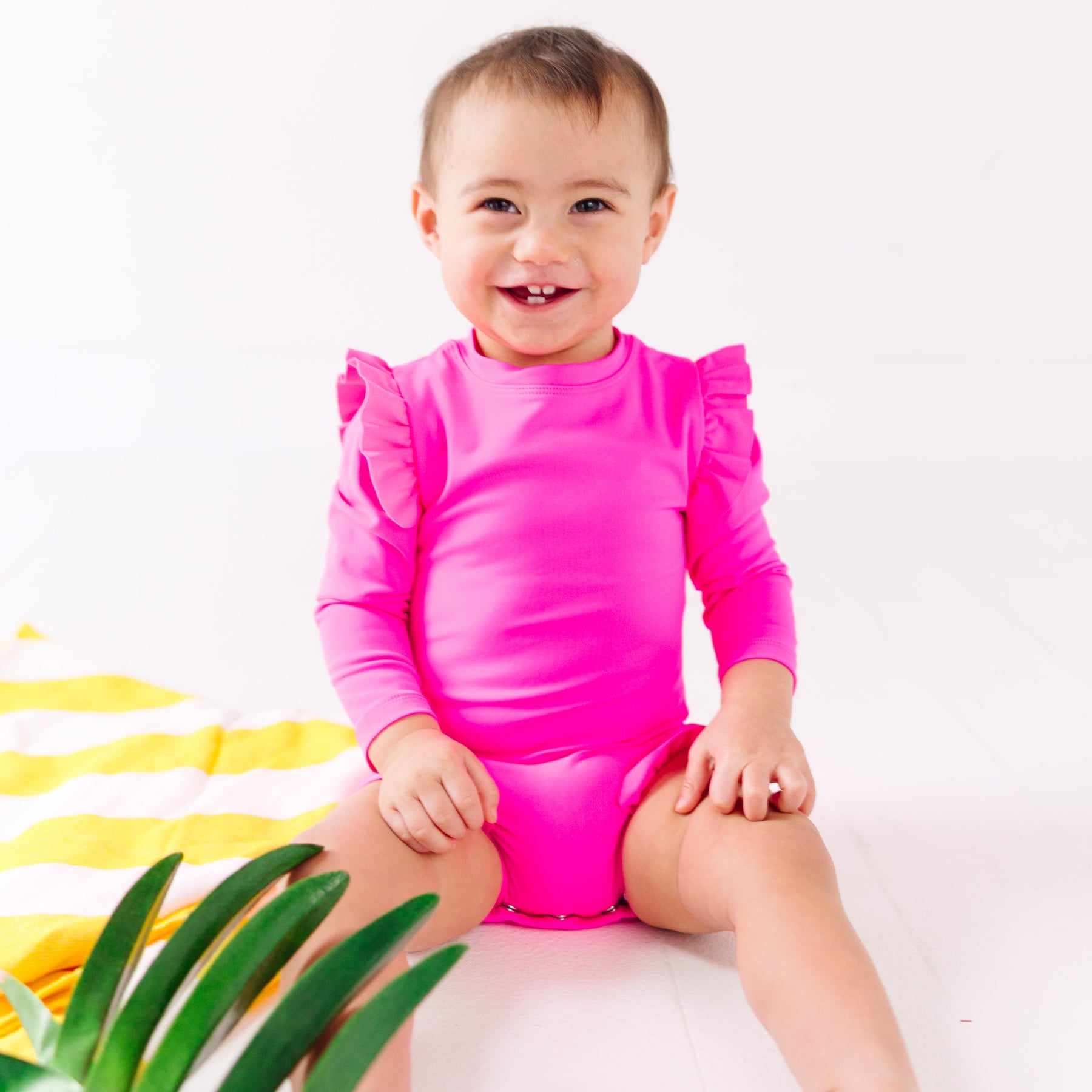 Baby wearing a bright pink one-piece swimsuit sitting on a white background with a yellow towel and green leaves.