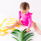 Baby wearing a bright pink one-piece swimsuit sitting on a white background with a yellow towel and green leaves.