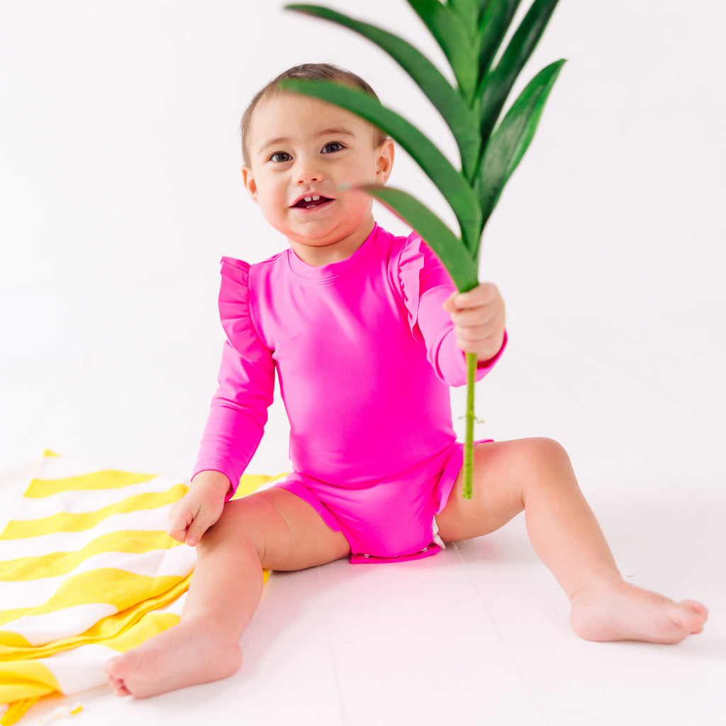 Baby wearing a bright pink one-piece swimsuit sitting on a white background with a yellow towel and holding green leaves.