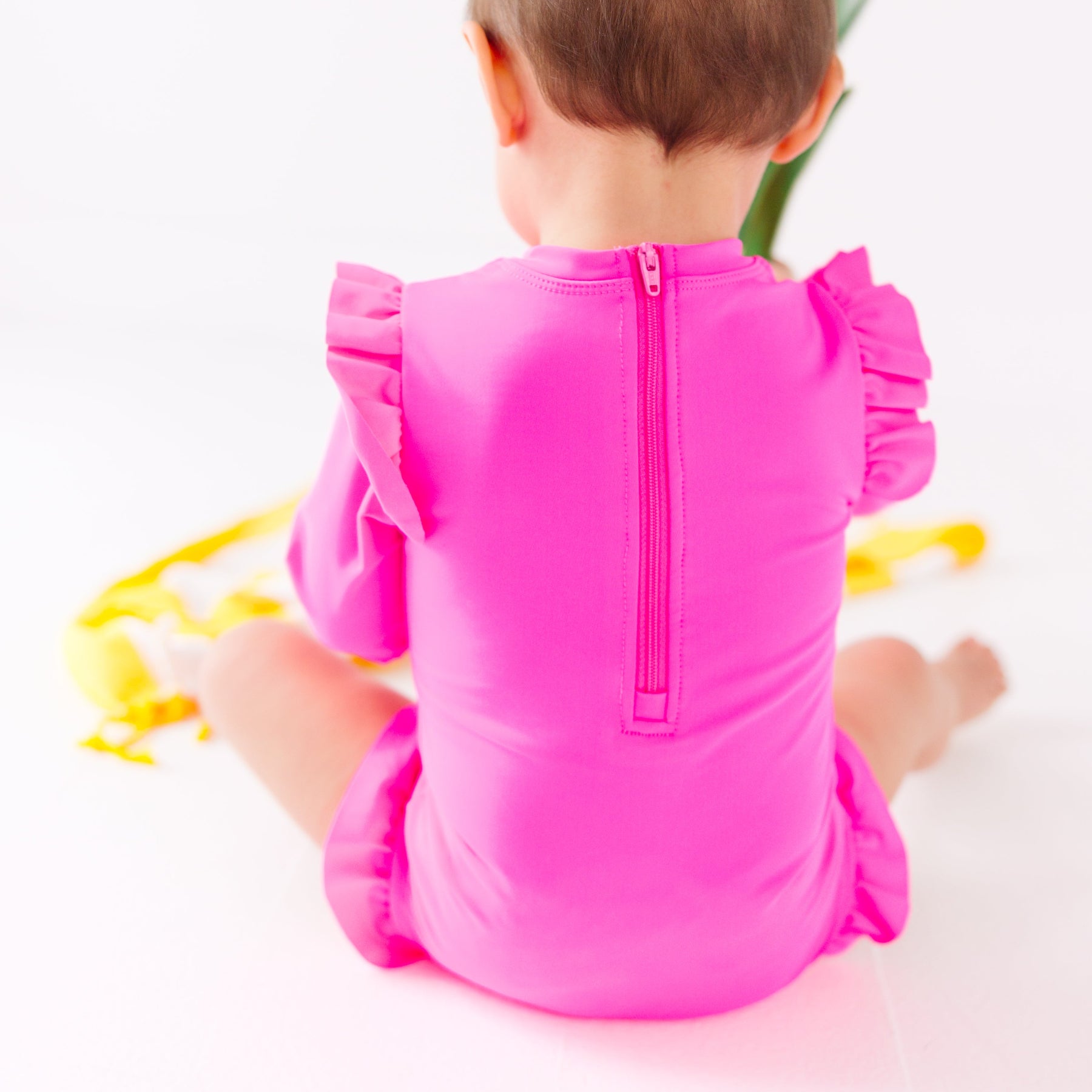 Back view of a baby wearing a bright pink one-piece swimsuit sitting on a white background with a yellow towel.