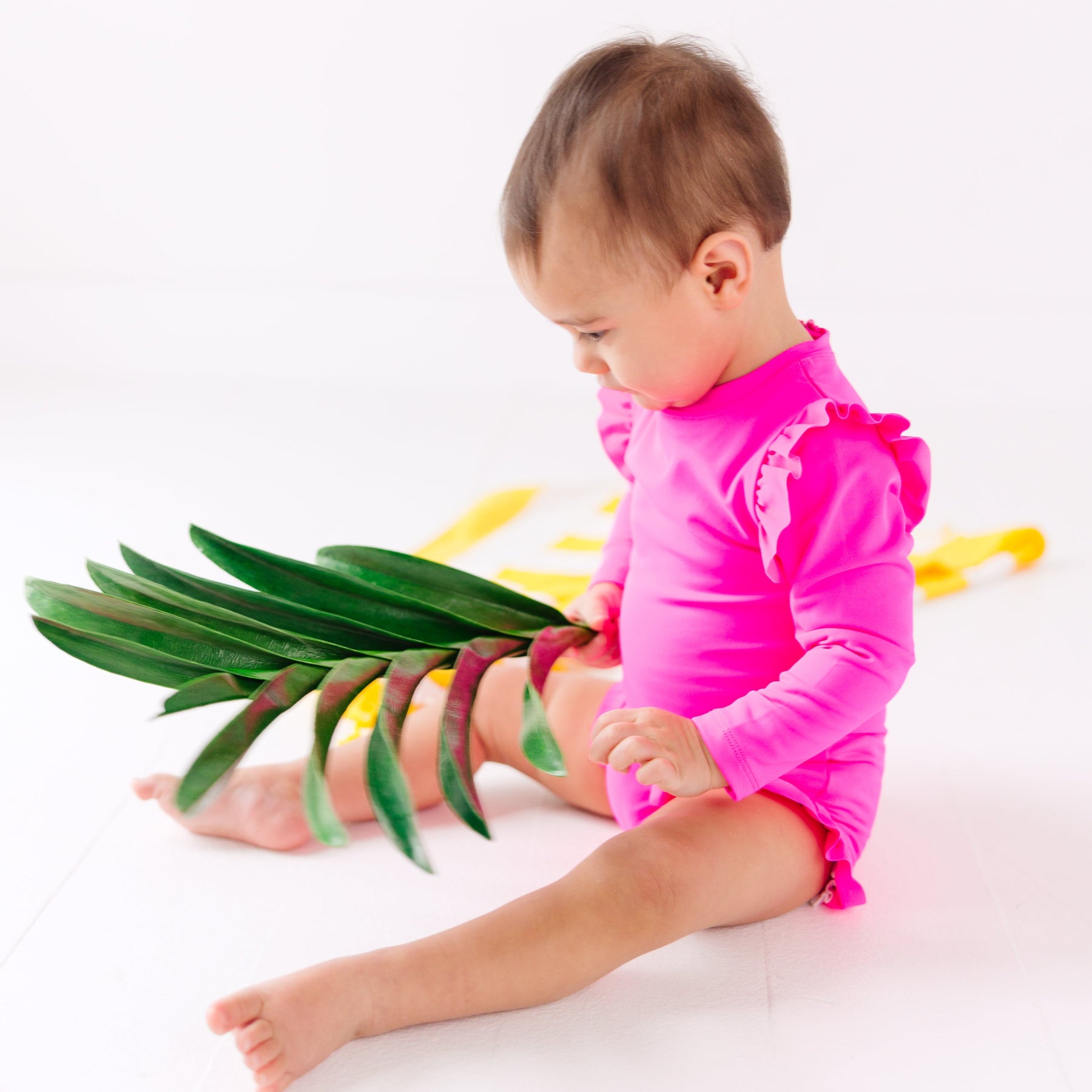 Baby wearing a bright pink one-piece swimsuit sitting on a white background with a yellow towel and holding green leaves.