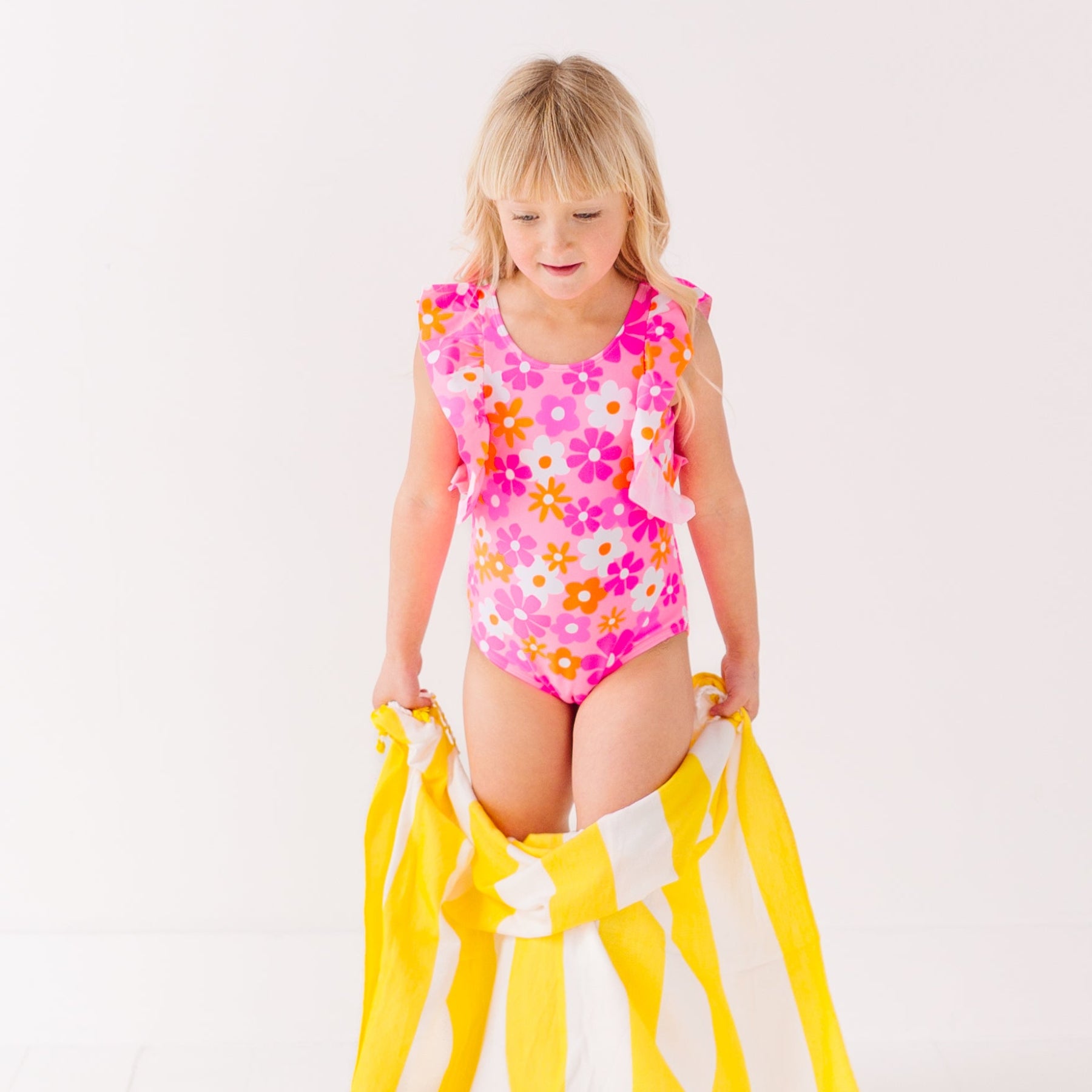 Child in a pink floral swimsuit with ruffles holding a yellow and white towel on a white background