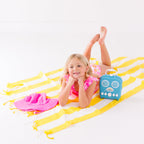 Child in a pink floral swimsuit with ruffles, lying on a yellow and white striped fabric with a blue radio toy and pink hat