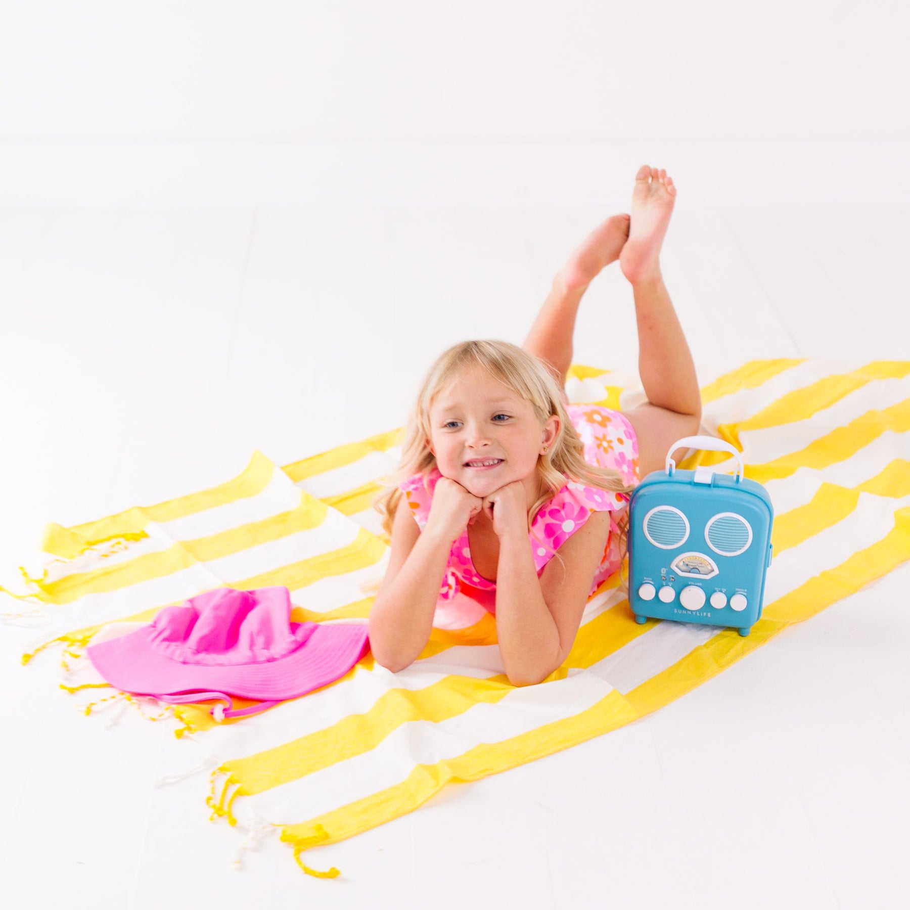 Child in a pink floral swimsuit with ruffles, lying on a yellow and white striped fabric with a blue radio toy and pink hat