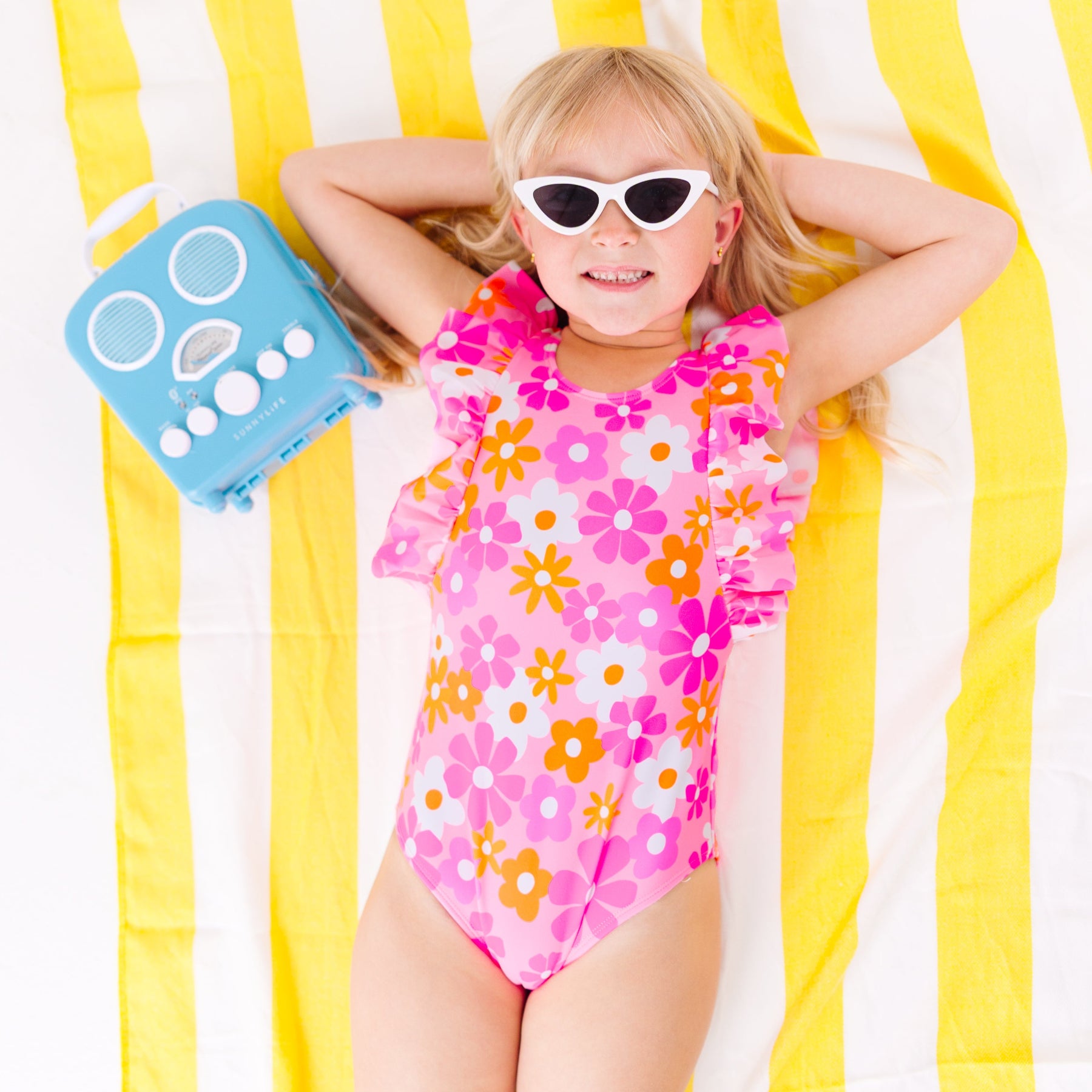 Child in a pink floral swimsuit with ruffles, wearing sunglasses, lying on a yellow and white striped fabric with a blue radio toy