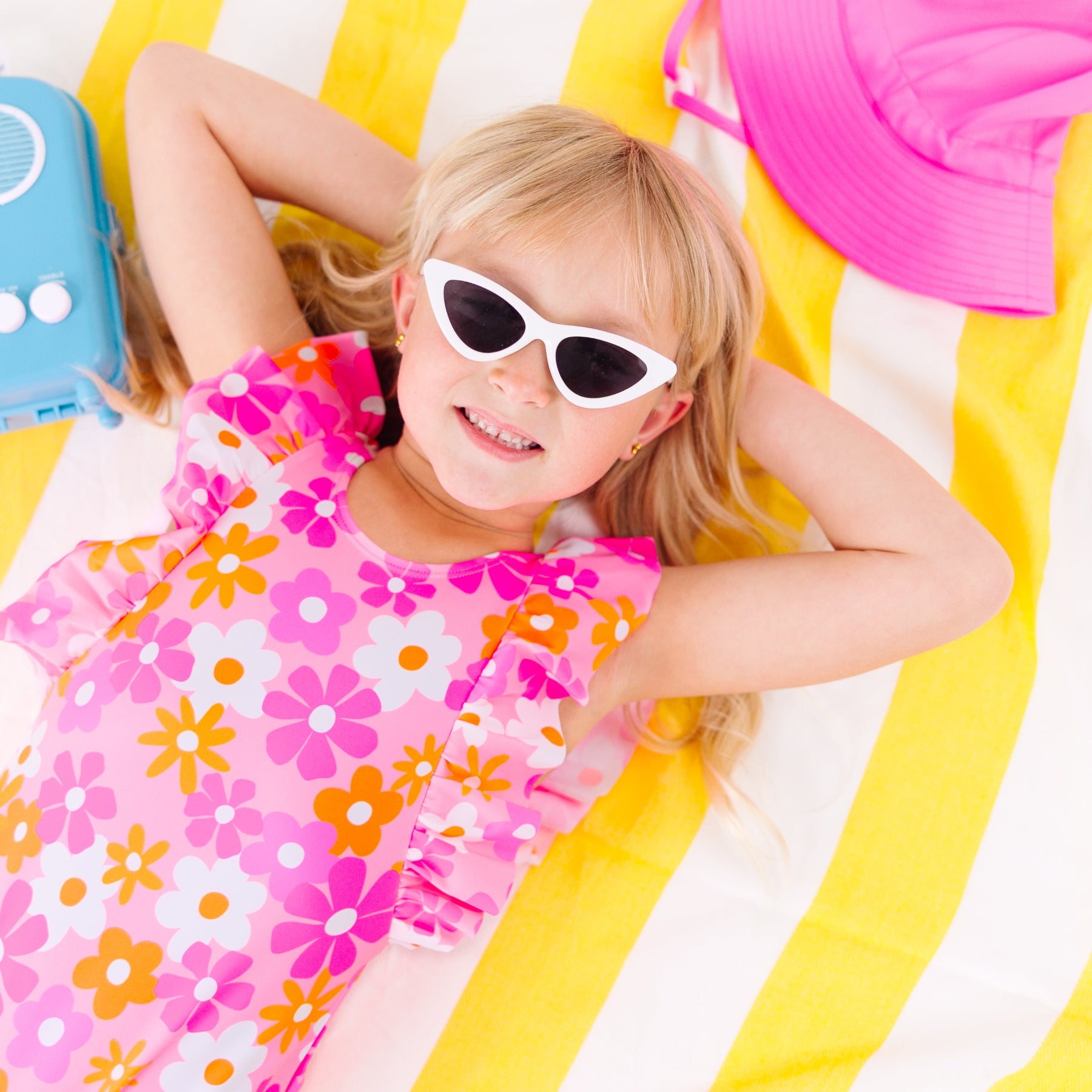 Child in a pink floral swimsuit with ruffles, wearing sunglasses, lying on a yellow and white striped fabric with a blue radio toy and pink hat