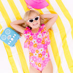 Child in a pink floral swimsuit with ruffles, wearing sunglasses, lying on a yellow and white striped fabric with a blue radio toy and pink hat