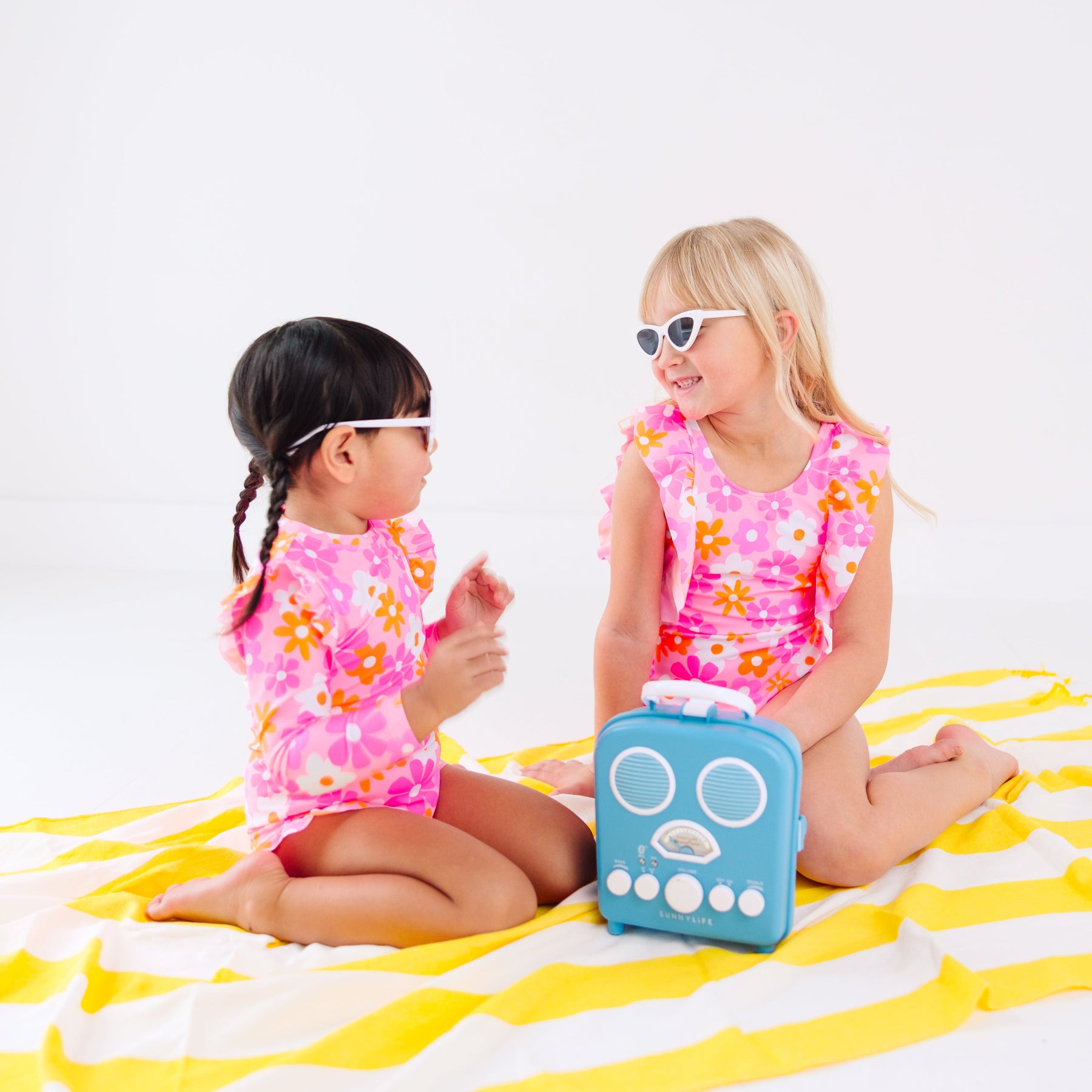 Two children in pink floral swimsuits sitting on a yellow and white striped towel with a blue radio toy.