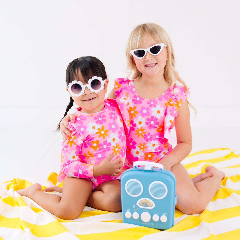 Two children in pink floral swimsuits sitting on a yellow and white striped towel with a blue radio toy.