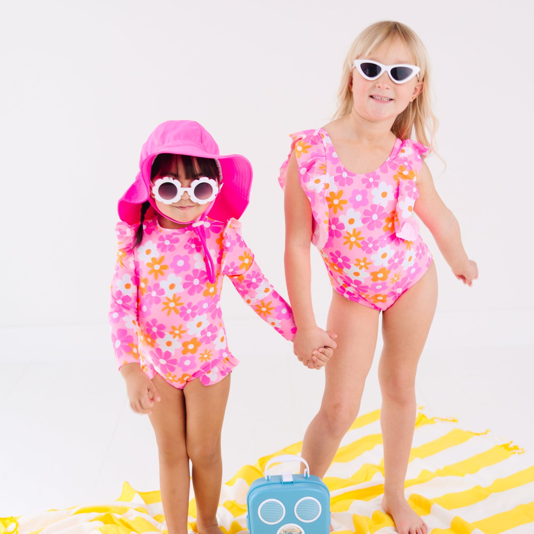Two children in pink floral swimsuits sitting on a yellow and white striped towel with a blue radio toy.