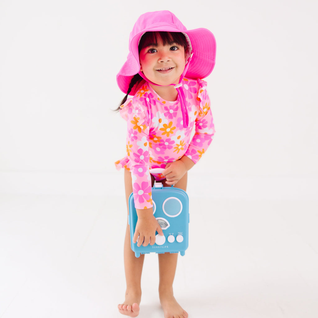 Child wearing a pink floral one-piece swimsuit and pink hat holding a blue radio toy on a white background