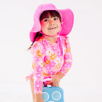 Child wearing a pink floral one-piece swimsuit and pink hat holding a blue radio toy on a white background