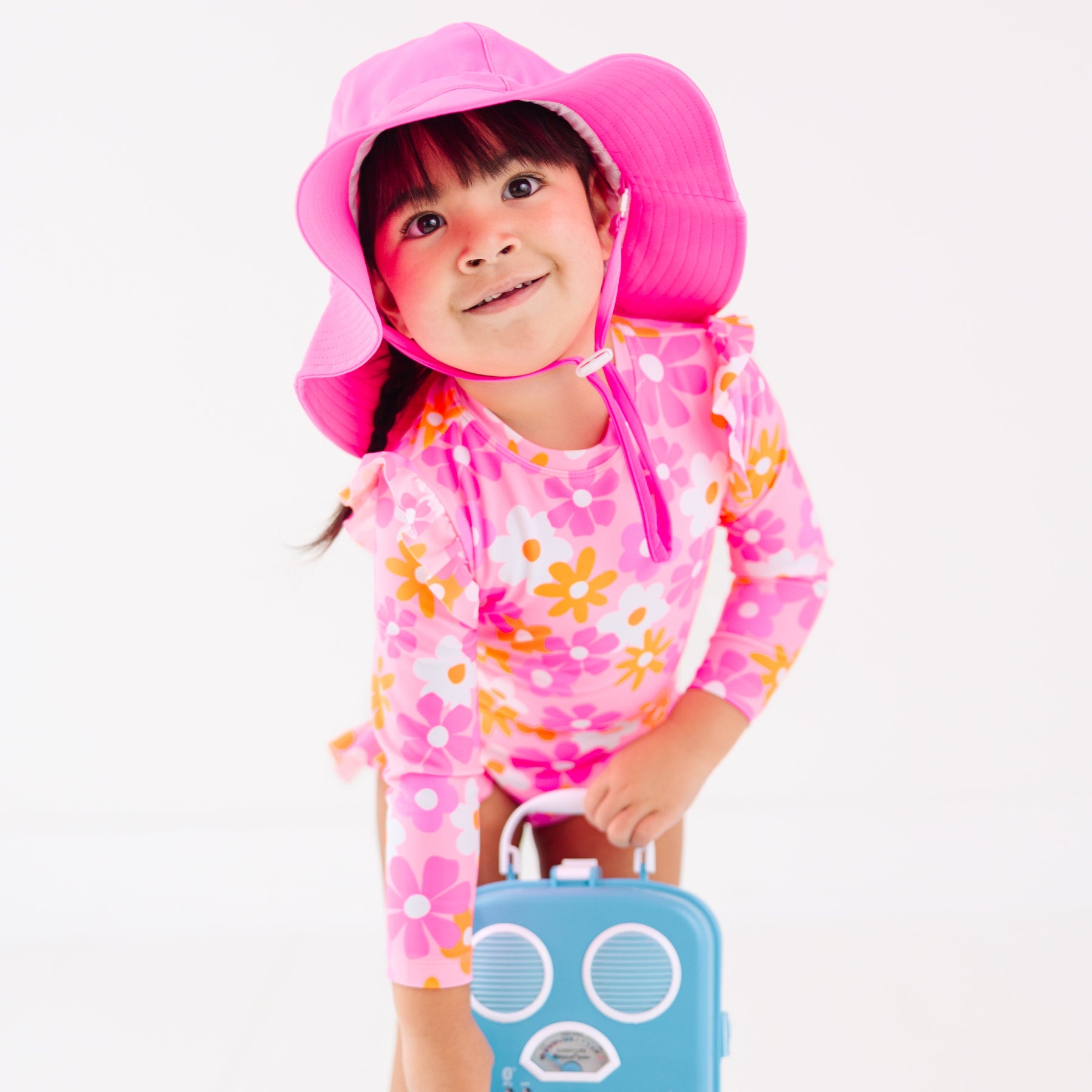 Child wearing a pink floral one-piece swimsuit and pink hat holding a blue radio toy on a white background
