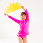 Child wearing a bright pink long sleeve two-piece swimsuit, holding a yellow sun-shaped object