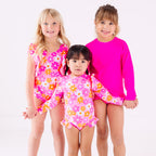 Three children wearing pink swimsuits with floral patterns on a white background