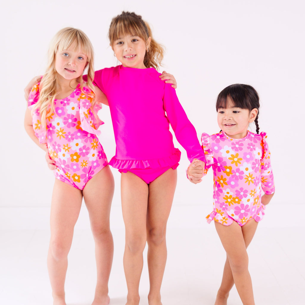 Three children wearing pink swimsuits with floral patterns on a white background