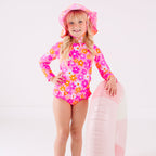 Child wearing a pink floral two-piece swimsuit and a matching sun hat while holding a pink floatie
