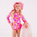 Child wearing a pink floral two-piece swimsuit and a matching sun hat while holding a pink floatie