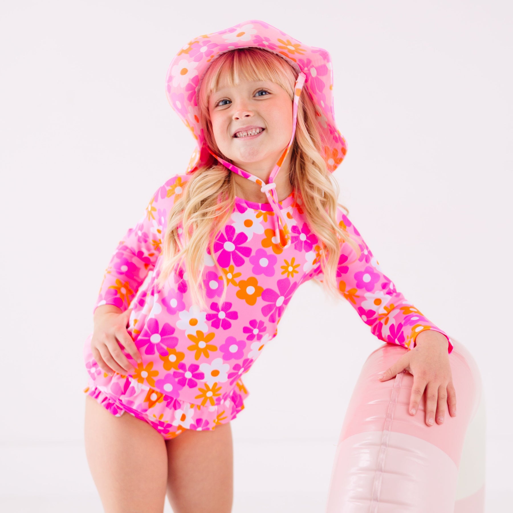 Child wearing a pink floral two-piece swimsuit and a matching sun hat while holding a pink floatie