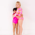 Two children in matching pink swimsuits standing on a white background