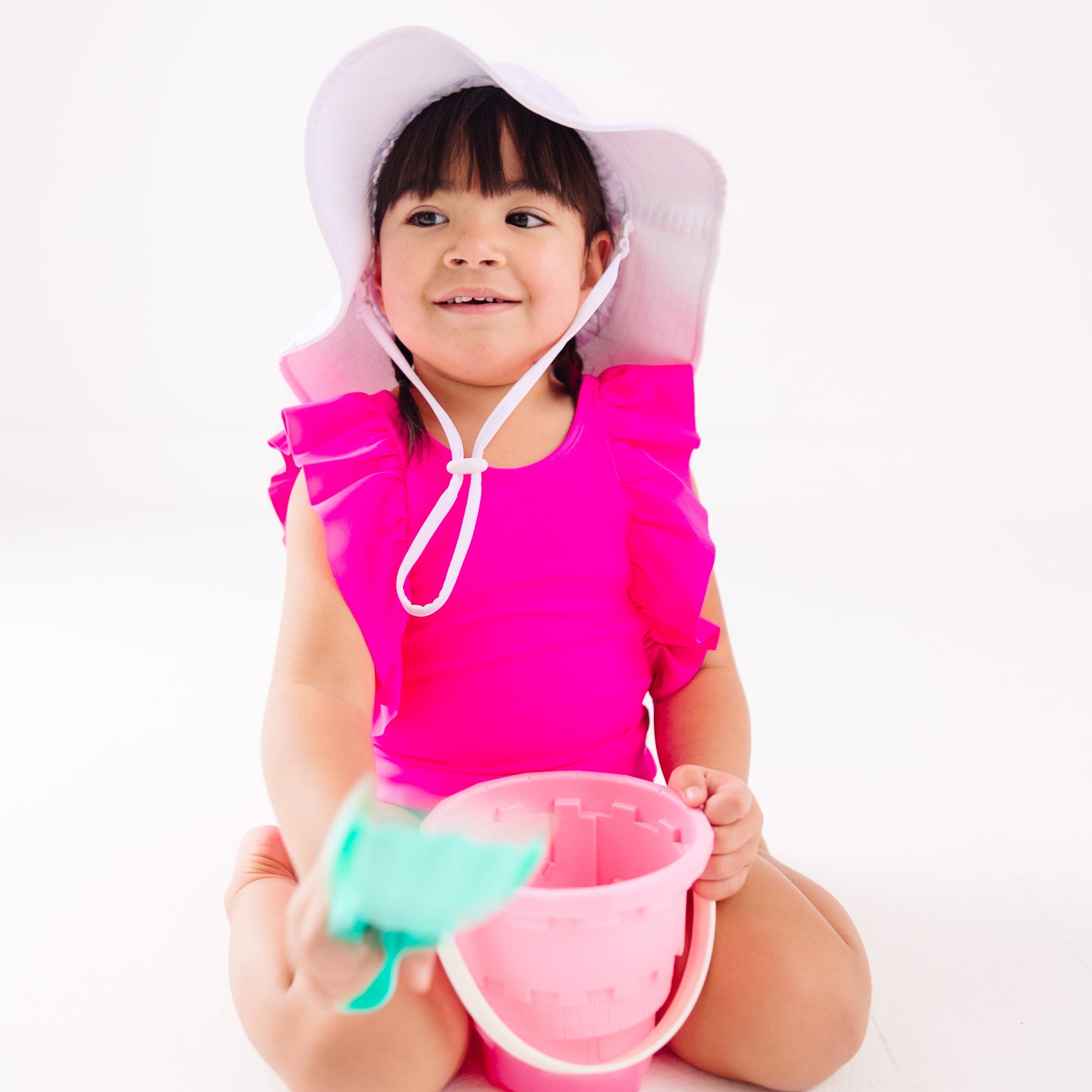 A young child in a bright pink swimsuit and white hat playing with a sand bucket and shovel on a white background