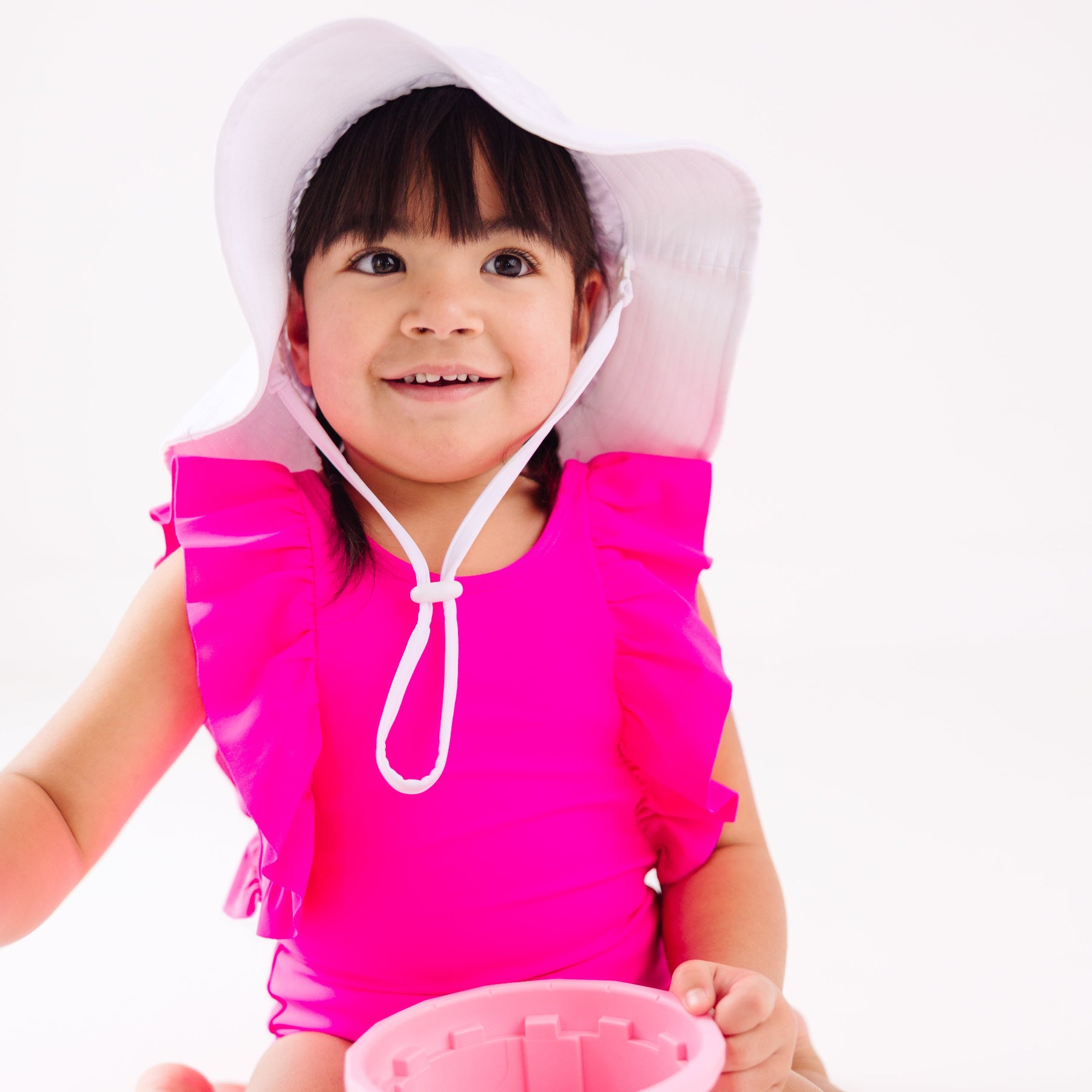 A young child in a bright pink swimsuit and white hat playing with a sand bucket on a white background