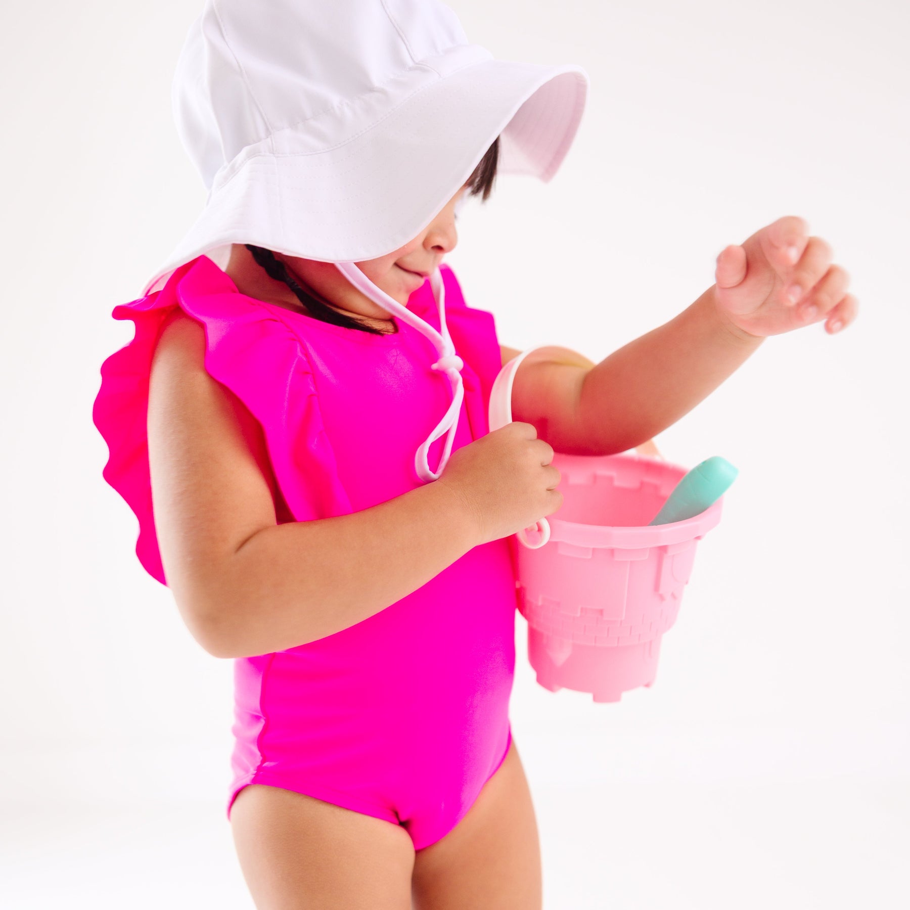 A young child in a bright pink swimsuit and white hat playing with a sand bucket on a white background