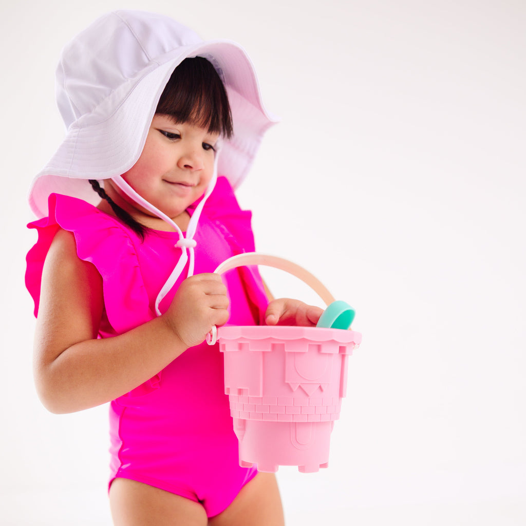A young child in a bright pink swimsuit and white hat playing with a sand bucket on a white background