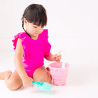 A young child in a bright pink swimsuit playing with a sand bucket and shovel on a white background