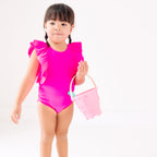 A young child wearing a bright pink swimsuit with ruffles holding a pink bucket on a white background