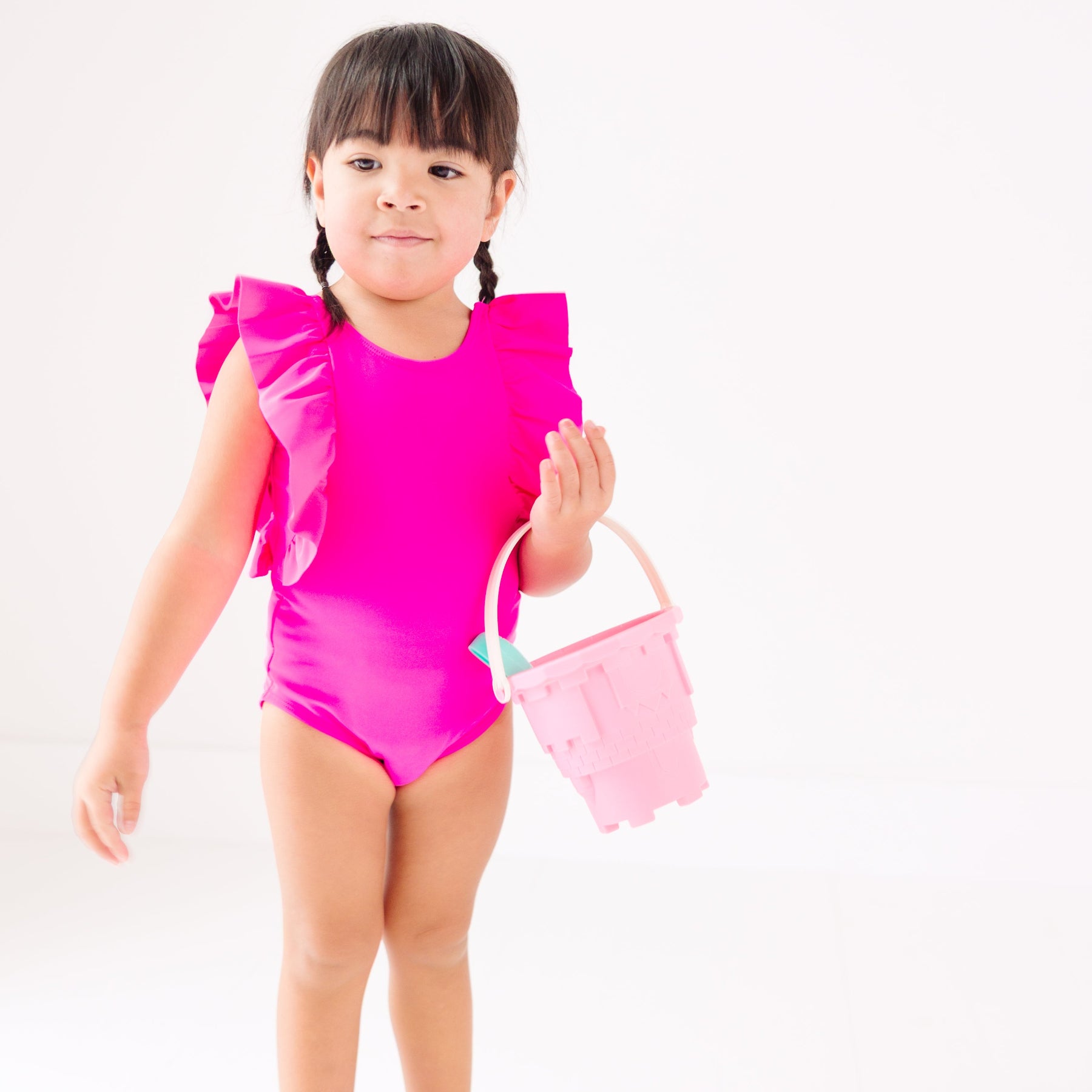 A young child wearing a bright pink swimsuit with ruffles holding a pink bucket on a white background