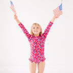 Young girl wearing a red floral swimsuit holding up two small American flags on a white background